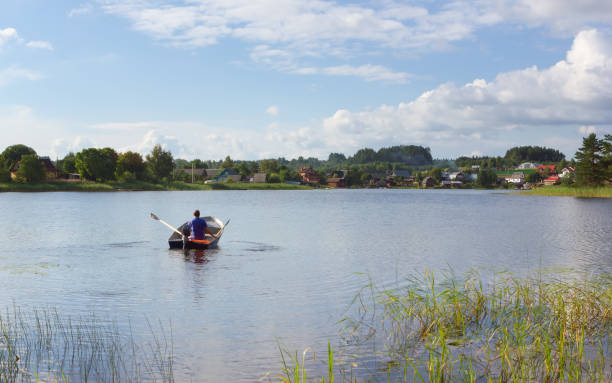 A man in a boat rowing to the village houses on a distant shore at sunny summer day. The concept of tourism, vacation, hobbies and outdoor activities - space for copy, selective focus. Lake Seliger, Russia.