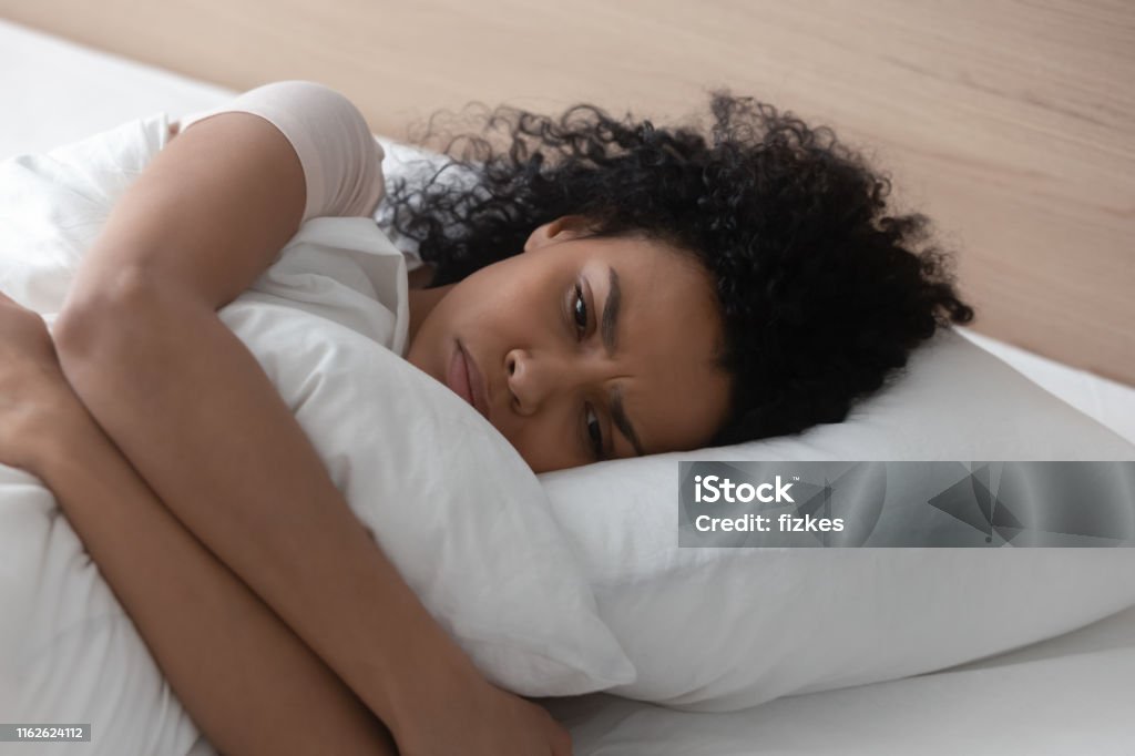 Sad depressed african woman hugging pillow lying in bed alone Sad depressed african american young woman hugging pillow lying in bed alone, upset frustrated black lady feeling lonely anxious suffer from insomnia trying to sleep thinking of problem in bedroom Women Stock Photo