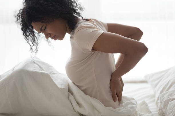 悲しい若いアフリカの女性は、ベッドの中で背中の痛みを感じて戻って触れる - human spine backache pain back ストックフォトと画像