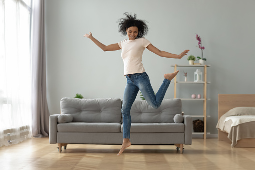 Carefree joyful african american millennial girl jumping dancing alone at home, happy funky active young fit mixed race woman having fun laughing flying in air in studio apartment room interior