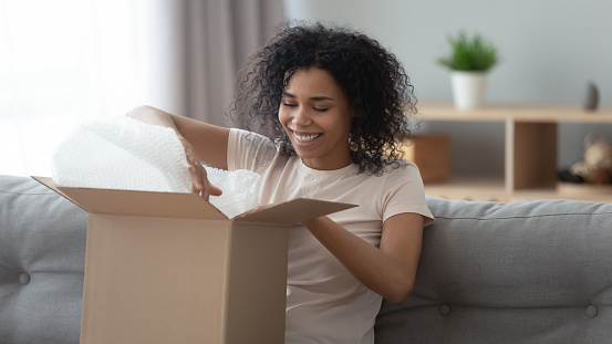Happy young african woman satisfied customer open parcel cardboard box sit on sofa at home, smiling black girl consumer unpack carton package receive good purchase postal shipping delivery concept
