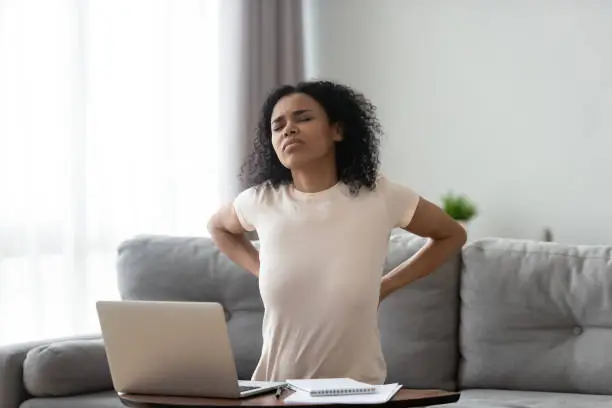 Young african woman feeling hurt ache muscle pain in back tired of computer sit in incorrect posture on sofa at home, black girl rubbing spine suffer from lower lumbar backache, backpain concept