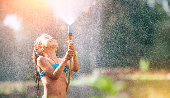 Cute little girl pours herself from the hose, makes a rain. Hot summer day pleasure