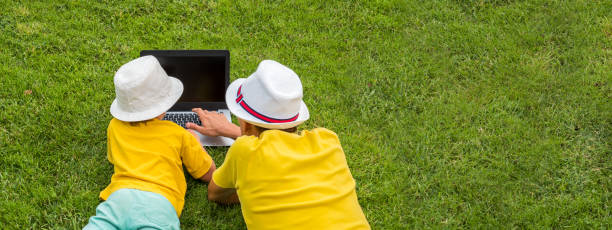 Father with son lying on the grass near laptop. Siblings and gadget outdoor. Father and son spending time together outdoor. Man and boy having fun on frontyard backyard. Gadget and children Copy space padre e hijo con cuaderno en el soleado día de verano. nuevo enfoque de la educación. vista trasera de dos chicos con computadora portátil en el patio trasero. feliz verano días soleados. niños con camisetas brillantes pasando tiempo al aire libre