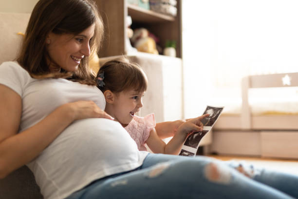 Cant wait to see my brother Pregnant mother cuddled up with daughter as they look at baby scan image together, smiling. Cute baby girl happy to see ultrasound image of her brother scientific imaging technique stock pictures, royalty-free photos & images