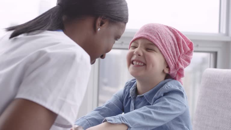 A black doctor sits on the floor with her patient. The patient is a caucasian girl with cancer. The girl is wearing a scarf on her head. The doctor is talking and holding the girl's hands. The child is smiling up at the doctor and listening intently.