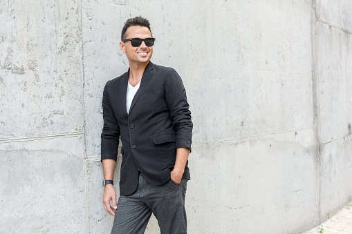 Young stylish man wearing suit and sunglasses outdoors on the city street isolated on concrete wall hand in pocket looking aside smiling cheerful