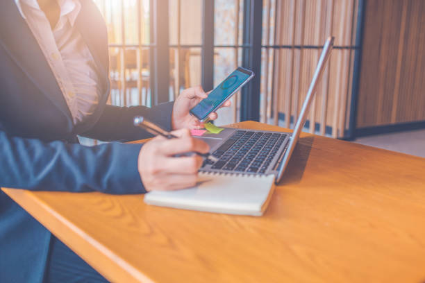 istock Business women are using mobile phones, screen displays work analysis charts, and she is taking notes on paper with a black pen in the office, a laptop computer placed on a wooden table in the office. 1162672684