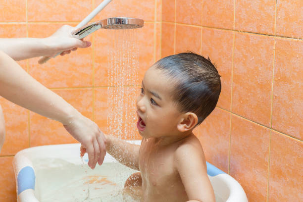 6-year-old boy squirts his face with the shower head during bath time in the bathtub.