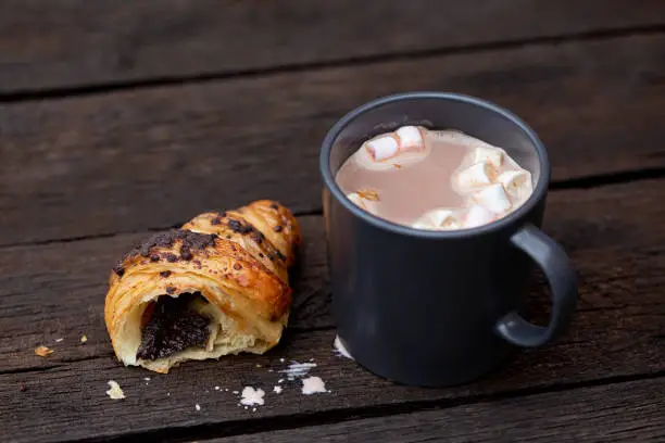 Hot chocolate with marshmallows in a blue-grey ceramic mug next to a half of chocolate croissant isolated on rustic dark brown wood table.