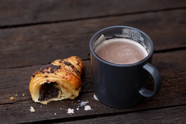 Hot chocolate in a blue-grey ceramic mug next to a half of chocolate croissant isolated on rustic dark brown wood table.
