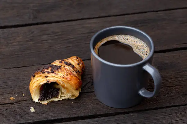 Black coffee in a blue-grey ceramic mug next to a half of chocolate croissant isolated on rustic dark brown wood table.