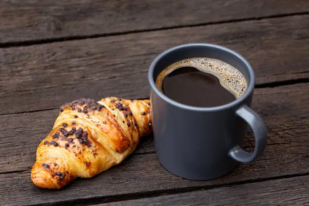 Black coffee in a blue-grey ceramic mug next to chocolate croissant isolated on rustic dark brown wood table.