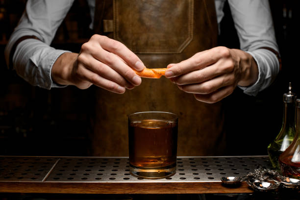 bartender añadiendo un jugo de ralladura de naranja a un cóctel de oro en el vaso con un gran cubo de hielo estampado - drink on top of ice food fotografías e imágenes de stock