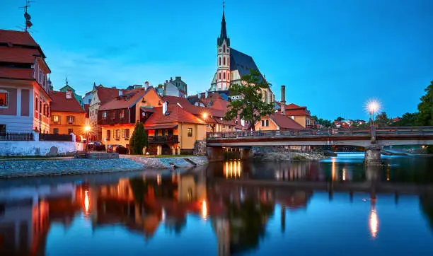Czech Krumlov, Czech Republic. Evening view at bridge in old town with old houses and broach at tower of St. Vitus Church. Sunset blue hour with illumination.
