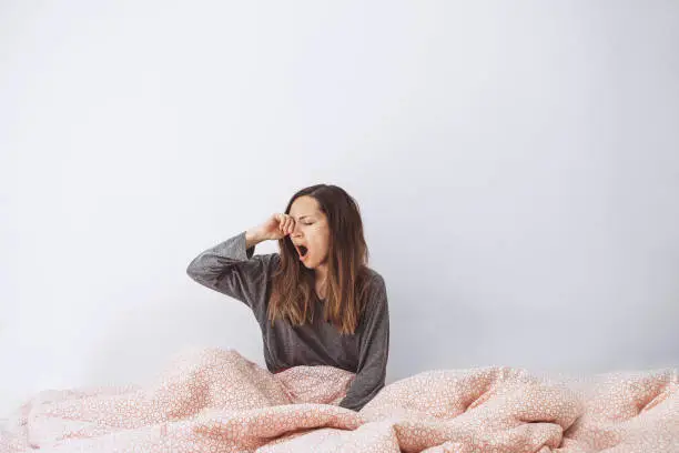 The girl is in bed and yawns. She covers her mouth with her hand. Early morning and she is sleepy.