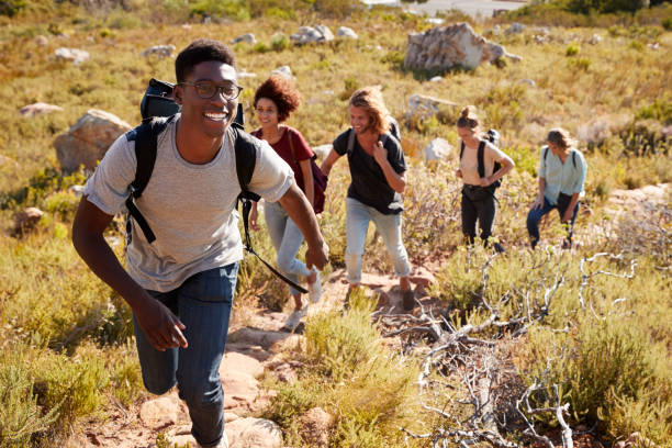 uomo afroamericano millenario che guida amici che si a piedi singolo file in salita su un sentiero in campagna - hiking foto e immagini stock