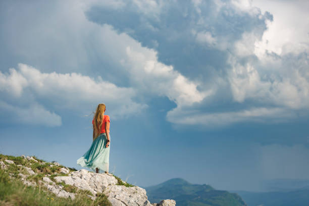 Selective focus on standing woman on top of the mountain viewed from behind unrecognisable person with storm and rain defocused in background.