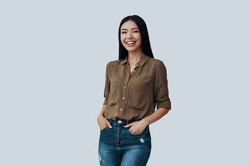 In good mood. Attractive young Asian woman looking at camera and smiling while standing against grey background
