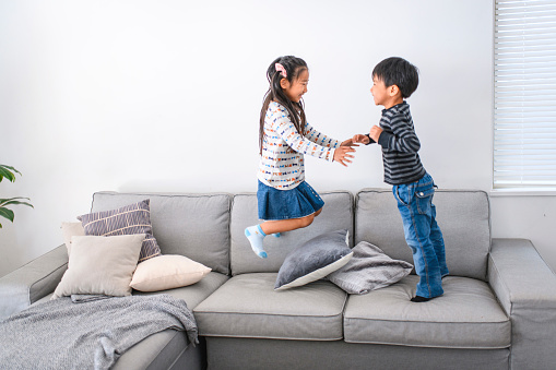 Cheerful young Japanese brother and sister jumping and playing on the living room sofa in their home.