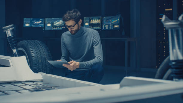 istock Automobile Design Engineer Sitting Beside Hybrid Electric Car Chassis Platform Prototype, Using Tablet Computer for Design Enhancement. Facility with Vehicle Frame with Suspension, Wheels, Engine and Battery 1162762309