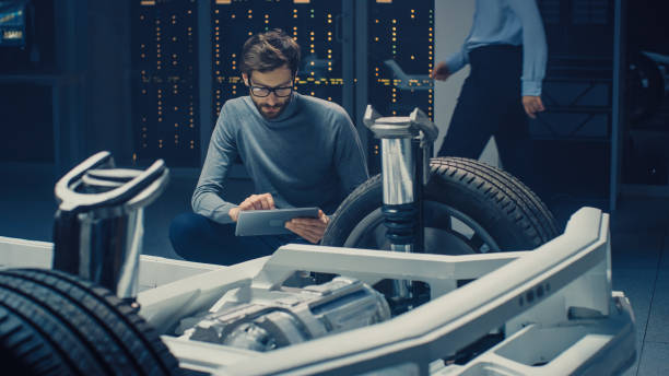 istock Automobile Design Engineer Sitting Beside Hybrid Electric Car Chassis Platform Prototype, Using Tablet Computer for Design Enhancement. Facility with Vehicle Frame with Suspension, Wheels, Engine and Battery 1162762402