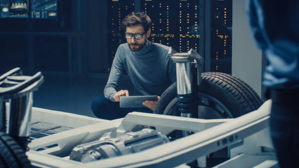 Automobile Design Engineer Sitting Beside Hybrid Electric Car Chassis Platform Prototype, Using Tablet Computer for Design Enhancement. Facility with Vehicle Frame with Suspension, Wheels, Engine and Battery