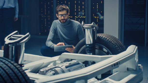 istock Automobile Design Engineer Sitting Beside Hybrid Electric Car Chassis Platform Prototype, Using Tablet Computer for Design Enhancement. Facility with Vehicle Frame with Suspension, Wheels, Engine and Battery 1162762465
