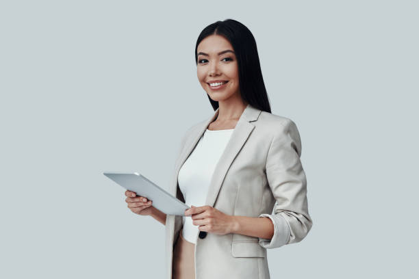 istock Young and smart. Attractive young Asian woman using digital tablet and smiling while standing against grey background 1162763617