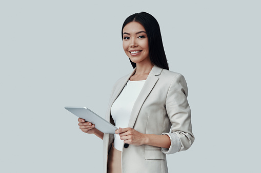 Young and smart. Attractive young Asian woman using digital tablet and smiling while standing against grey background