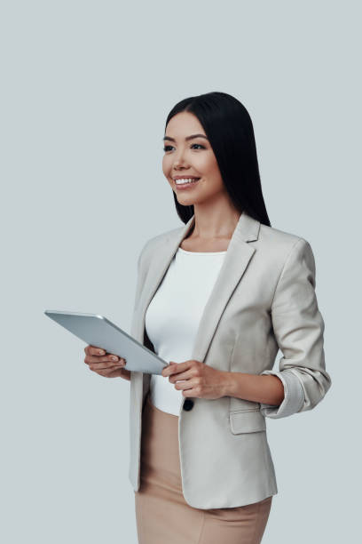 istock Always ready to help. Attractive young Asian woman using digital tablet and smiling while standing against grey background 1162763707