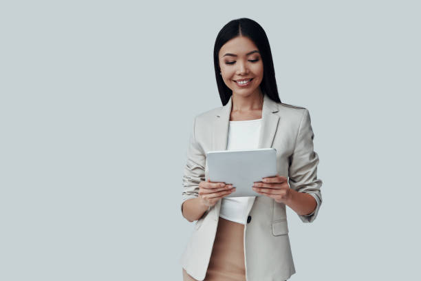 istock Confident expert. Attractive young Asian woman using digital tablet and smiling while standing against grey background 1162763843