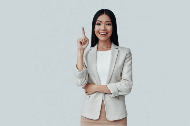 istock Take a look! Attractive young Asian woman looking at camera and pointing copy space while standing against grey background 1162766073