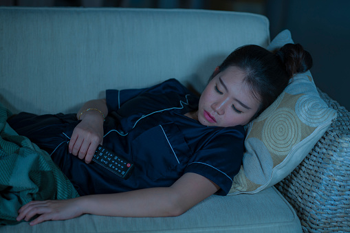 home lifestyle portrait of young beautiful and tired Asian Korean woman in pajamas holding TV remote falling asleep on living room sofa couch while watching television show or movie at night