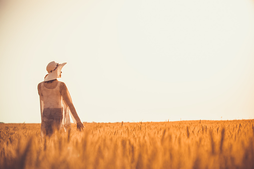 Copy space shot of happy young woman in sunhat standing in endless golden fields of barley and enjoying summer sunshine while looking away.