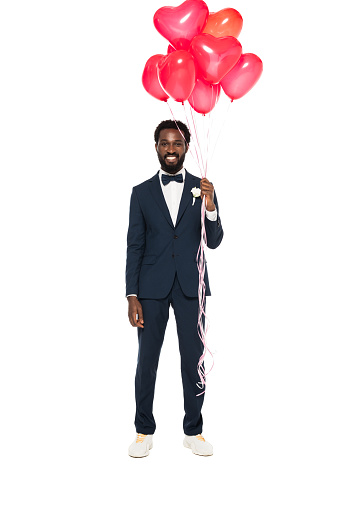 handsome african american bridegroom holding pink heart-shaped balloons and standing isolated on white
