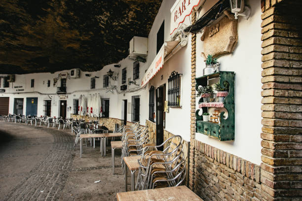 View along the buildings built into the rocky cliff in Setenil de las Bodegas, Andalusia, Spain with empty restaurant tables and chairs curving away