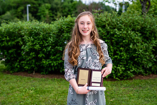 Young teen girl/Middle school student standing in front of a bush/garden after her graduation ceremony while holding a diploma and academic awards.