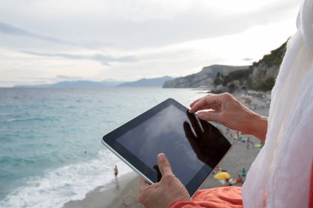 woman uses digital tablet while sitting on a wall above the sea - beach stone wall one person imagens e fotografias de stock