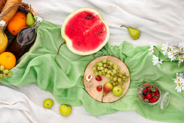 istock Summer picnic in the meadow on the green grass. Fruit basket, juice and bottled wine, watermelon, strawberries in a glass jar and baguettes of bread. White tablecloth and a bouquet of field daisies. 1162802425