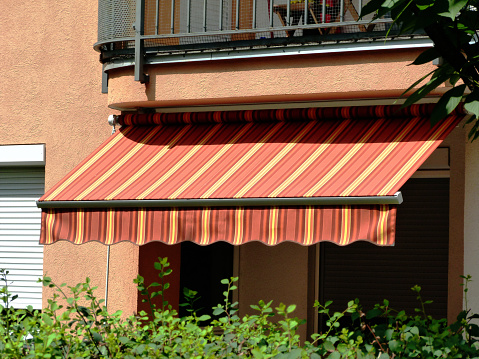colorful canvas awning in yellow & orange color above a residential balcony. sunny summer day. sun protection concept. UV exposure protection. warm color waterproof fabric material. green garden. selective focus.