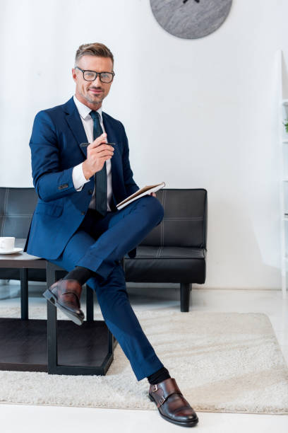 istock cheerful businessman in glasses holding notebook and pen while sitting on coffee table with crossed legs 1162815795