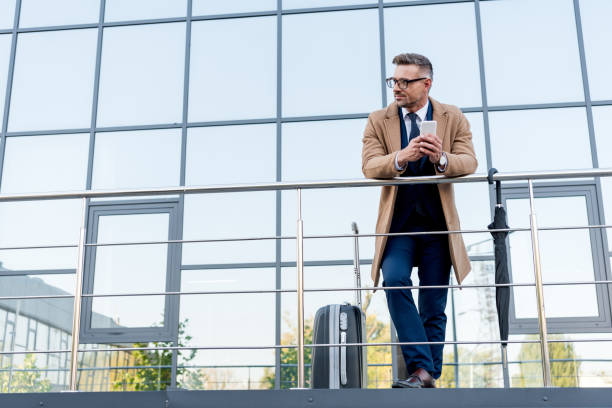 istock cheerful businessman in beige coat using smartphone while standing near suitcase and umbrella 1162815805