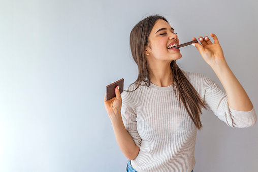 Young woman with natural make up having fun and eating chocolate isolated on gray background. Woman eating chocolate. Happy teenage girl eating chocolate bar