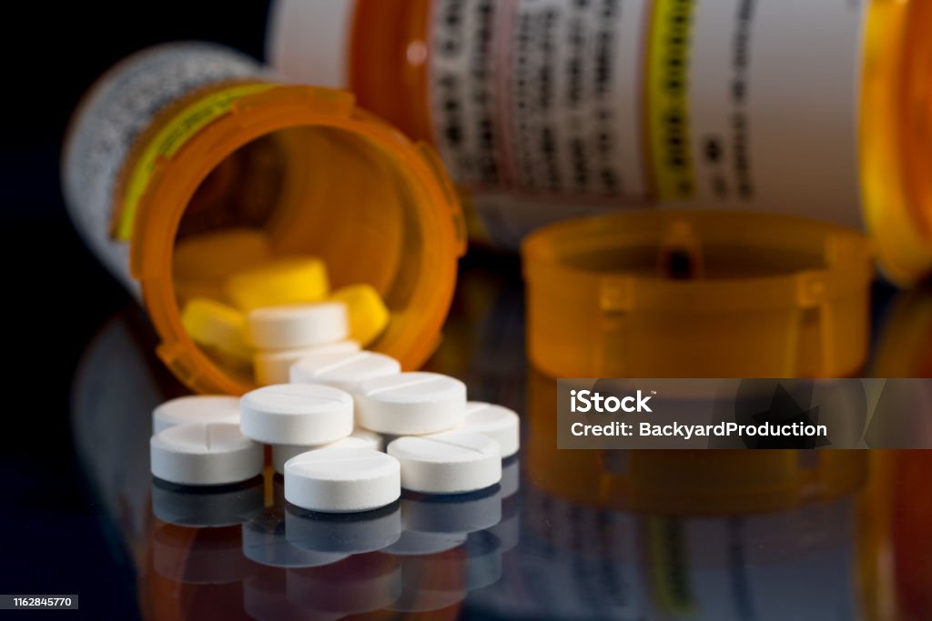 Macro of oxycodone opioid tablets with prescription bottles against dark background Oxycodone is the generic name for a range of opoid pain killing tablets. Prescription bottle for Oxycodone tablets and pills on glass table with reflections Medicine Stock Photo