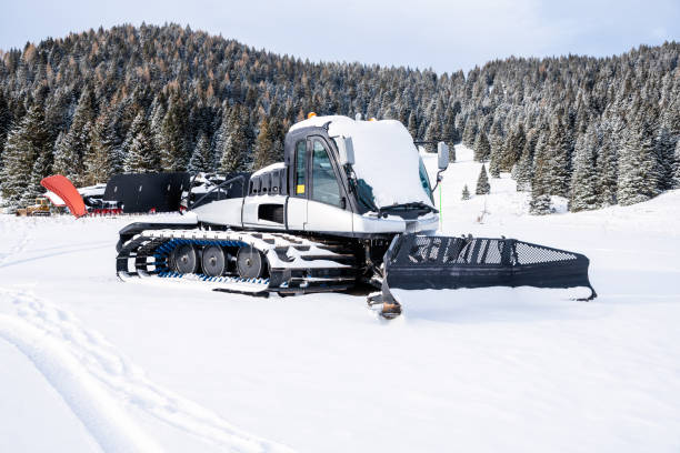 Snow groomers meticulously smooth and prepare ski slopes in a snowy mountain landscape under clear skies and bright sunlight, ensuring optimal skiing conditions.