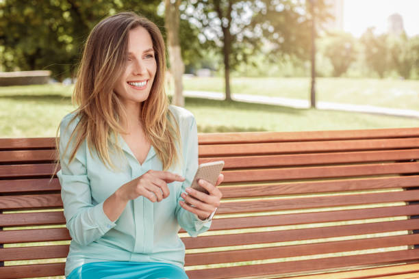 istock Young woman sitting on bench touching screen playing game on smartphone laughing joyful 1162865297