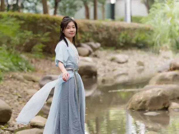 Beautiful Asian woman in cyan Chinese costume clothes hanfu beside of stream and looking at camera in peaceful smile, traditional ancient Chinese beauty.