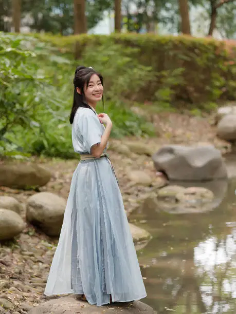 Beautiful Asian woman in cyan Chinese costume clothes hanfu beside of stream and looking at camera in peaceful smile, traditional ancient Chinese beauty.