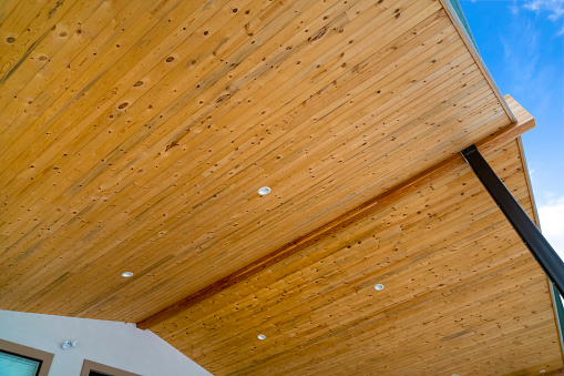 Close up view of the underside of the roof of a house with blue sky background. Round lights are installed on the ceiling made of brown wooden planks.